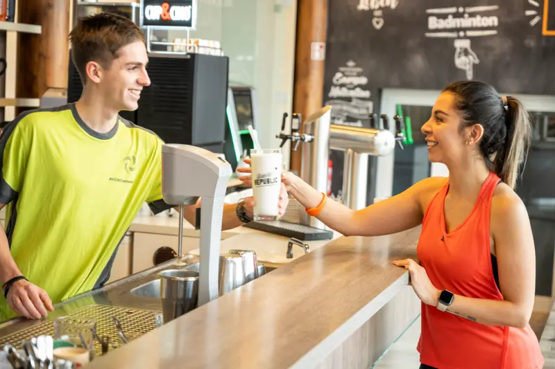 Ein Mitarbeiter reicht einer lächelnden Sportlerin am Check-in im Fitnessbereich ein frisches Getränk
