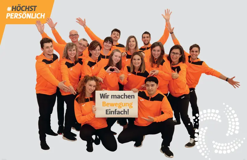 Das Team des Sportpark Johannesplatz in orangen Jacken mit ausgebreiteten Armen und Herzen, dazu das Schild: Wir machen Bewegung Einfach!