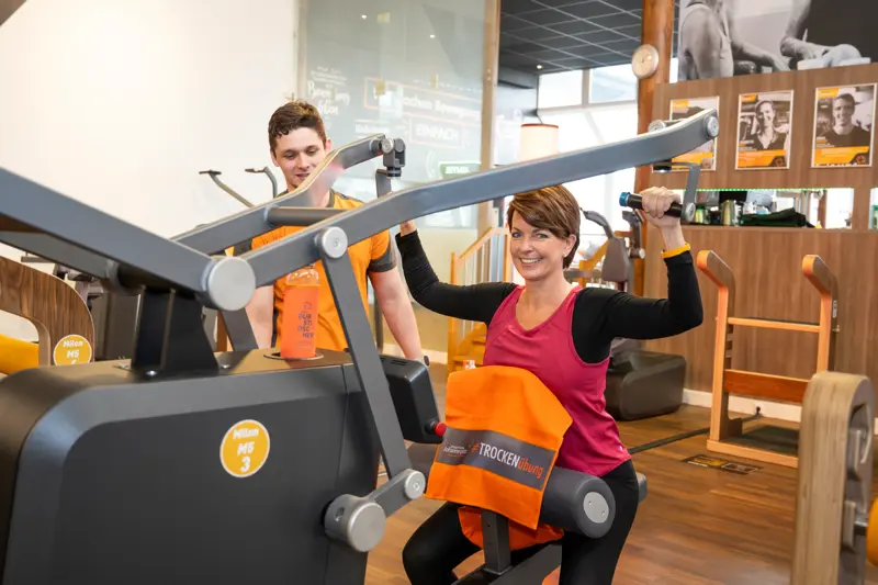 Eine Frau trainiert an einem Milon-Gerät, ein Trainer steht freundlich hinter ihr